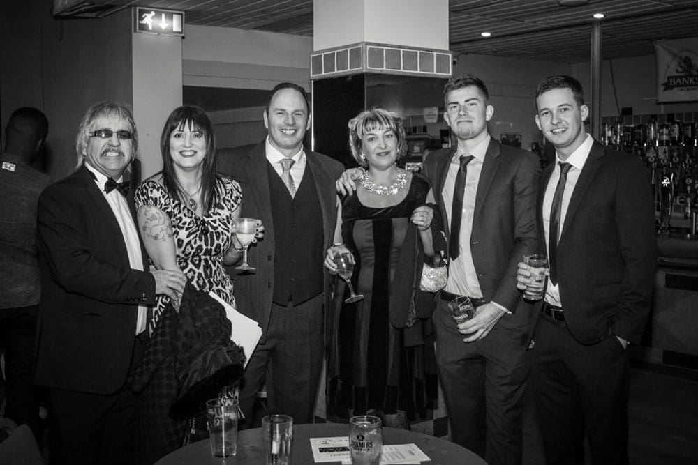 A group of six adults dressed formally, smiling and holding drinks, pose together in a bar or lounge setting. Captured by a Birmingham event photographer, the photo is in black and white.