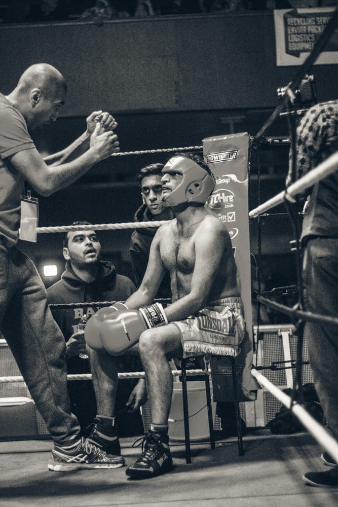 A boxer wearing gloves and headgear sits in his corner as his coach gives instructions; two team members watch nearby inside the boxing ring, skilfully captured by a Birmingham event photographer.