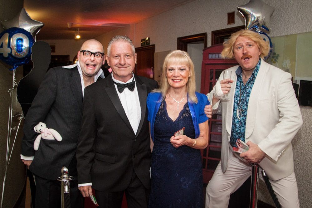 Four adults dressed in formal and party attire pose together at an event with balloons and a red telephone box in the background. Captured by a Birmingham event photographer, one man wears a wig and holds a drink. All are smiling at the camera.